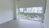This contemporary single-floor space embodies a clean, minimalist design philosophy that prioritizes light and openness. The most striking architectural feature is the wide, horizontal band of windows that spans a full wall, offering unobstructed views of a tree-lined urban streetscape and the surrounding modern buildings. These oversized windows flood the interior with abundant natural light, creating a bright and airy atmosphere throughout.

The flooring is finished in light-toned hardwood, contributing to the space's fresh and modern character while adding a sense of warmth to the otherwise neutral palette. The walls are rendered in crisp white, complementing the natural light and enhancing the sense of spaciousness.

The open layout provides a versatile and uncluttered environment, well-suited for a variety of uses. The seamless connection between the interior and the urban exterior, facilitated by the expansive glazing, gives the space a dynamic and connected feel. Overall, this space offers a refined, contemporary environment defined by its thoughtful use of natural light, quality finishes, and a streamlined design that balances simplicity with sophistication.