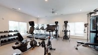 The fitness center at Parkside At The Beach is a thoughtfully designed, single-floor facility that provides a comprehensive workout environment. The space features crisp white walls, neutral carpeted flooring, and recessed lighting complemented by ceiling fans, creating a clean and comfortable atmosphere conducive to exercise.

The cardio zone is well-stocked with Matrix treadmills, elliptical trainers, and stationary and cycling bikes, all equipped with individual display screens. Multiple large windows fitted with white blinds line the walls, allowing abundant natural light to fill the space while offering views of the surrounding greenery.

The strength training area features a multi-functional cable machine system, a Smith machine with adjustable benches, and a full rack of dumbbells organized on tiered storage. Large wall mirrors expand the sense of space and allow users to monitor their form during workouts. A wall-mounted flat-screen television is positioned for visibility across the strength training zone.

Additional functional equipment, including stability balls and resistance tools, rounds out the facility's offerings. The layout is spacious and open, allowing for comfortable movement between equipment. Access to the gym is provided through both interior and glass-paneled exterior doors.