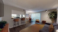 This single-floor residence offers a thoughtfully designed layout that balances comfort and functionality. The open-plan living and dining area forms the heart of the home, where wood-look flooring, a textured ceiling, and neutral walls create a warm, inviting atmosphere. The living room features a wall-mounted flat-screen TV, a cozy sofa, and stylish décor accents including bold artwork and lush greenery, complemented by a contemporary swing-arm wall sconce. The dining area is anchored by a round wooden table beneath a pendant light, with a full-height sliding glass door dressed in vertical blinds that floods the space with natural light and offers a view to the outdoors. The galley-style kitchen is efficiently organized with white cabinetry, dark granite-look countertops, and black appliances including a refrigerator, electric range, and dishwasher, all set over wood-look flooring. The bedroom is a restful retreat highlighted by a deep navy accent wall, warm wood flooring, a walk-in closet, and a bright window that brings in natural light. The full bathroom is finished with a dark countertop vanity, a large mirror, and a tub-shower combination, completing this well-rounded living space.