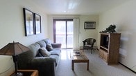 This single-floor space offers a welcoming and functional layout centered around a comfortable living area. The living room is furnished with a plush gray sofa, a wooden glass-top coffee table, and a wicker accent chair, creating a relaxed and inviting atmosphere. A wall-mounted entertainment unit anchors one side of the room, while framed artwork adds a personal touch to the walls. Natural light pours in through a large sliding glass door fitted with vertical blinds, which opens directly onto a private balcony — an ideal spot to enjoy outdoor views of the surrounding landscape. A wall-mounted air conditioning unit ensures year-round climate comfort. The carpeted flooring adds warmth underfoot and contributes to the overall cozy ambiance of the space. The neutral color palette throughout keeps the interior feeling clean, open, and easy to personalize. Whether used as a primary residence or a getaway retreat, this space combines practical living features with a relaxed, comfortable character suited to everyday use.