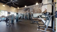 This single-floor fitness center is designed to support a full range of workout styles within a spacious, open layout. The facility is divided into distinct functional zones, each tailored to a specific type of training. The strength training area is anchored by a striking exposed brick accent wall featuring bold motivational murals — including the phrases "Break Your Limits" and "Don't Quit, Get Fit" — creating an energetic, industrial-inspired atmosphere. This zone is equipped with cable machines, adjustable benches, and weight racks, with large mirrors lining one wall to maximize visual space and assist with form.

A dedicated functional training area offers TRX suspension straps mounted to the wall, kettlebells, medicine balls, and stability balls organized on a TAG-branded rack. Rubber flooring throughout provides durability and impact absorption across all training zones.

The cardio section features a row of treadmills and elliptical machines positioned in front of expansive floor-to-ceiling windows that offer natural light and outdoor views of palm trees, giving the space an open, airy feel. Mounted flat-screen televisions accompany the cardio equipment for entertainment.

The main open area features soaring ceilings with exposed dark ceilings, pendant lighting, and ceiling fans, contributing to an industrial-modern aesthetic. The overall design balances functionality with visual appeal.