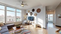 This contemporary single-floor residence offers a well-designed layout that balances communal living with private retreats. The open-concept kitchen is a standout feature, equipped with sleek, high-gloss blue-grey cabinetry, stainless steel appliances, a subway tile backsplash, and a generous peninsula with bar seating — ideal for casual dining and entertaining. Light wood-style flooring flows throughout the main living areas, complementing the bright, airy atmosphere created by an abundance of natural light.

The living room impresses with tall windows that frame outdoor views of the surrounding neighborhood, a ceiling fan, and a vibrant teal accent wall adorned with colorful artwork. The space comfortably accommodates a large sectional sofa and separate seating area, making it both functional and inviting.

The bedrooms are thoughtfully appointed, with large windows — some floor-to-ceiling — that flood the rooms with daylight and offer urban views. Each bedroom features neutral tones, carpeted flooring, and ample space for a study area.

The bathrooms are finished in a clean, modern style with grey vanity cabinetry, white countertops, large mirrors, and glass-enclosed walk-in showers with white tile surrounds. Each bathroom maintains a consistent, contemporary aesthetic that ties the overall design of the residence together.
