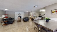 This single-floor space offers a functional and welcoming open-concept layout designed for group gatherings and shared use. The living area serves as the centerpiece of the space, highlighted by a dramatic floor-to-ceiling white stone fireplace wall accented with decorative brick detailing. Comfortable leather seating and round side tables create a relaxed lounge atmosphere throughout this area.

The fully equipped kitchen runs along a galley-style layout, featuring gray cabinetry with ornate detailing, light stone countertops, a tile backsplash, stainless steel appliances including a refrigerator and electric range, a built-in microwave, and ample storage. The kitchen opens directly into the main living and dining areas for easy flow.

Two distinct dining areas provide generous seating capacity, each furnished with sturdy wood tables and chairs. One dining space benefits from abundant natural light through multiple windows fitted with horizontal blinds, while painted brick walls and a central entry door add a grounded, classic character to the room.

The accessible bathroom is thoughtfully appointed with a wall-mounted sink, a large framed mirror with vanity lighting, tile wainscoting, and ADA-compliant grab bars. Light tile flooring runs consistently throughout the entire space, tying the rooms together with a clean, neutral aesthetic.