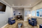Located at 575 McDonough Pkwy in McDonough, GA, this thoughtfully designed lounge space offers a polished and inviting atmosphere within a compact, single-room footprint. The interior is finished with warm neutral-toned walls accented by soft blue paint, complemented by crisp white crown molding that frames the ceiling throughout. Recessed lighting and a decorative glass pendant fixture provide layered illumination, enhancing the room's welcoming ambiance.

The space is furnished with bold cobalt blue leather accent chairs equipped with attached side tables, arranged to encourage conversation and comfortable use. A low rectangular coffee table anchors the central seating area, while a wall-mounted flat-screen television serves as a focal point on one wall. A cozy dining nook tucked into an alcove features a dark wood table and bench seating beneath the pendant light, adding functional versatility to the room.

Patterned carpet tiles in earthy tones of tan and brown ground the space and add visual texture underfoot. Curated abstract artwork in coordinating blue and neutral hues adorns the walls, contributing to a cohesive, contemporary aesthetic. A window along one wall admits natural light and provides views of the surrounding building exterior. A set of double doors with an illuminated exit sign offers direct access to adjacent areas, completing this well-considered, multi-purpose lounge space.