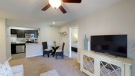 This single-floor residence offers a thoughtfully designed open-concept layout that seamlessly connects the main living spaces. The living area is anchored by plush wall-to-wall carpeting and features a ceiling fan with a light fixture, contributing to a comfortable and airy atmosphere. A white media console with lattice-detail cabinetry provides functional storage and display space, while a decorative coastal wall sculpture adds a touch of character to the neutral walls.

Adjacent to the living area, a cozy dining space accommodates a round glass-top table paired with wicker-style chairs, creating an inviting spot for casual meals. The kitchen is visible from the dining area and showcases dark cabinetry contrasted against light countertops, stainless steel appliances, and a tiled backsplash, delivering both style and practicality.

A doorway from the living area leads directly into the bedroom, which features soft carpeting and a calm, restful ambiance consistent with the rest of the home. Throughout the residence, the cohesive color palette of warm beiges and soft whites creates a bright, welcoming environment. Recessed lighting and ample natural light further enhance the open and comfortable feel of this well-designed space.