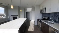 This single-floor space showcases a contemporary open-concept design with a well-appointed kitchen at its core. The kitchen features dark espresso cabinetry paired with a striking white quartz island countertop, creating a bold contrast that anchors the space. Stainless steel appliances, including a range, built-in microwave, and smooth cooktop, are seamlessly integrated alongside a dark subway tile backsplash that adds texture and visual depth. A pendant light hangs above the island, providing focused task lighting while contributing to the modern aesthetic. Wide-plank hardwood-style flooring in a warm grey-brown tone runs throughout, unifying the space. Natural light filters in from multiple directions, highlighting the clean lines and neutral grey wall palette. Adjacent to the kitchen, a fireplace with a dark surround offers a cozy focal point, suggesting a connected living area. A hallway leads further into the space, hinting at additional rooms beyond. The overall ambiance is clean, modern, and functional, with thoughtful design details that balance style and everyday practicality.