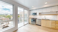 This single-floor unit offers a thoughtfully designed layout that maximizes functionality within a compact footprint. Light hardwood flooring runs consistently throughout, contributing to a cohesive, modern aesthetic anchored by a clean white color palette.

The kitchen serves as a central feature, outfitted with stainless steel appliances including a gas range, microwave, and dishwasher. Two-tone cabinetry combines white upper cabinets with warm natural wood lower cabinets, complemented by a geometric tile backsplash and a undermount sink. A large sliding glass door floods the kitchen with natural light and provides direct access to an outdoor deck with open views.

The hallways are efficiently organized, with one housing a stacked washer and dryer unit behind a sliding barn door, maximizing space without sacrificing convenience. A built-in floor-to-ceiling wood storage unit with open shelving and closed cabinetry provides ample organizational capacity in an adjacent area.

The bathroom features a full-size soaking tub with a tub-surround enclosure and chrome fixtures, finished in a clean, minimal white. Recessed lighting throughout the unit ensures consistent illumination, reinforcing the bright, airy character of the overall space.