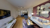 This single-floor residence showcases a thoughtfully designed open-concept layout that connects the living, dining, and kitchen areas into one cohesive and functional space. The living area is anchored by a bold deep-burgundy accent wall adorned with decorative metallic circular mirrors, creating a striking focal point. A white sofa accented with colorful throw pillows, a modern lounge chair, and a gold-toned coffee table furnish the space, all resting atop a vibrant purple and grey area rug over warm honey-toned hardwood flooring.

The kitchen is finished with white upper cabinetry, dark lower cabinetry, and stainless steel appliances, including a refrigerator and range. A sleek kitchen island with bar seating provides additional functionality and serves as a natural dividing element between the cooking and living zones. Track lighting runs along the ceiling, supplementing recessed lighting throughout to ensure the space is well-illuminated.

The overall ambiance is contemporary and vibrant, blending clean architectural lines with bold design choices. Light-colored walls and smooth ceilings contribute to a bright, airy feel, while the cohesive color palette of purples, whites, and warm wood tones ties the entire space together elegantly.