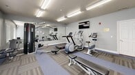 Located at 882 W Foxboro Dr in North Salt Lake, Utah, this dedicated fitness center offers a well-organized and fully equipped exercise space designed to support a well-rounded workout routine. The room is finished with neutral gray walls, patterned modular carpet tiles, and bright overhead fluorescent lighting that keeps the space feeling open and energized.

A large wall-length mirror along one side visually expands the room and allows users to monitor their form during exercise. Equipment includes a multi-station cable machine for strength training, a Star Trac treadmill, an elliptical trainer, a stationary bike, a rowing machine, and a weight bench. A tiered dumbbell rack stocked with hex weights provides free weight options for a variety of exercises.

A wall-mounted flat-screen television adds an entertainment element for those looking to stay engaged during cardio sessions. Two windows dressed with horizontal blinds allow for natural light and ventilation, while hand sanitizer is conveniently mounted on the wall for hygiene. Motivational "inhale" and "exhale" framed signs add a welcoming, wellness-oriented touch to the décor. The space is cleanly maintained and thoughtfully arranged to maximize functionality within its footprint.