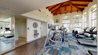 Located in Chandler, Arizona, this fitness center offers a spacious and well-appointed workout environment spread across a single floor. The facility's most striking architectural feature is its dramatic vaulted ceiling with rich, exposed wood beams that rise to impressive heights, creating an open and energizing atmosphere. Tall, arched windows line the walls, flooding the interior with natural light and providing views of the surrounding landscaped grounds.

The main workout area is carpeted in a modern blue and gray patterned flooring, housing a comprehensive lineup of Life Fitness cardio equipment, including treadmills, ellipticals, and stationary bikes arranged for easy access. A dedicated strength training section features a cable machine, weight bench, and a squat/power rack loaded with plate weights. A flat-screen TV is mounted for entertainment during workouts.

A secondary area with hardwood-style flooring includes a full-length mirrored wall, ideal for stretching or functional training, and provides convenient access to the entry. Additional amenities include a wall-mounted water fountain, a wall clock, and restroom access. The combination of high ceilings, ample natural light, quality equipment, and thoughtful layout makes this fitness center a functional and inviting space for a full range of workout routines.