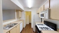 This single-story space showcases a well-designed galley-style kitchen as its central feature. The kitchen is laid out in a narrow, elongated configuration with countertops and cabinetry lining both sides, maximizing the use of available space. Light wood-toned cabinets line both the upper and lower sections of the walls, providing generous storage throughout. The countertops offer practical workspace, complemented by a stainless steel sink positioned along one side.

The kitchen is fully equipped with white appliances, including a gas range with a black cooktop, a built-in over-the-range microwave, a full-sized refrigerator, and a dishwasher, creating a cohesive and functional cooking environment. Overhead, a flush-mounted fluorescent light fixture provides bright, even illumination throughout the space.

The flooring features dark hardwood-style planks that run the length of the kitchen, adding warmth and contrast to the light cabinetry and white walls. At the far end of the kitchen, a doorway opens into an adjoining room with yellow-toned walls, suggesting additional living or utility space beyond. The overall ambiance is clean, practical, and efficiently organized.
