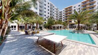 This outdoor amenity space offers a resort-inspired experience defined by its generous pool deck, vibrant tropical landscaping, and thoughtfully designed recreational zones. At its heart lies a large, shimmering pool with crystal-clear blue water, surrounded by paver decking and an array of lounge seating and shaded cabanas, creating an inviting environment for relaxation and socializing. Tall palm trees are strategically placed throughout the courtyard, providing natural shade and lending a distinctly tropical ambiance to the space.

The surrounding walkways are finished in high-quality interlocking pavers that wind through lush garden beds filled with tropical foliage, including palms, ornamental shrubs, and ground-level plantings that add layers of color and texture. Recreational amenities include a dedicated ping-pong table positioned poolside, encouraging an active and social atmosphere.

Elevated walkways with sleek glass railings frame portions of the space, offering elevated vantage points overlooking the pool and gardens below. Large glass-paned doors and floor-to-ceiling windows on the ground-level building facades connect interior spaces to the outdoors, maximizing natural light and visual continuity. The crisp white architectural exterior of the surrounding buildings, accented with warm yellow balcony detailing, complements the bright, open-air setting — creating a cohesive and upscale outdoor living environment.
