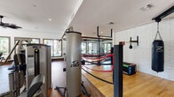 This comprehensive fitness center is thoughtfully organized into distinct workout zones, each designed to support a full range of exercise disciplines. The cardio area features a row of modern treadmills positioned in front of large, dark-framed windows that frame lush outdoor views of palm trees and green landscaping, filling the space with abundant natural light. Elegant curtain accents and hardwood-style flooring add a refined touch to the room.

The strength training zone is generously appointed with Cybex selectorized machines, a full rack of dumbbells, kettlebells, weight benches, and cable systems. Floor-to-ceiling mirrors line the walls, enhancing the sense of space, while mounted flat-screen TVs and a bold ceiling fan overhead contribute to an energizing atmosphere.

A standout feature of the facility is its dedicated boxing area, complete with a full boxing ring defined by red ropes, hanging heavy bags, a speed bag station, and whitewashed brick walls that lend an industrial character. Polished hardwood floors and glass panel dividers give this zone a sleek, professional feel.

The group fitness studio offers light hardwood floors, spin bikes, stability balls, medicine balls, yoga mats, and BOSU trainers, all bathed in natural light from expansive windows. Dual ceiling fans and a large wall mirror complete this versatile studio, making it suitable for a wide variety of classes and individual training sessions.