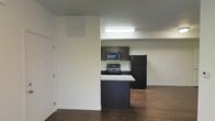 This single-floor space features an open-concept layout where the kitchen flows seamlessly into the adjacent living area, creating a cohesive and functional environment. The kitchen is equipped with dark espresso-toned cabinetry that provides ample storage, complemented by a full suite of black appliances, including a gas range, over-the-range microwave, and a full-size refrigerator. A peninsula counter with a light-colored surface extends from the cabinetry, offering additional prep space and serving as a natural divider between the kitchen and the rest of the living area. The backsplash behind the range adds a subtle design detail to the cooking zone. Overhead, a flush fluorescent light fixture provides direct task lighting over the kitchen workspace. The flooring throughout is a dark hardwood-style material that grounds the space with a warm, rich tone and contrasts effectively with the crisp white walls. The neutral wall color keeps the interior feeling clean and bright. A wall-mounted HVAC vent is visible near the entry, indicating a central climate control system. The overall design is practical and straightforward.