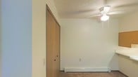 This single-floor residential space offers a straightforward and functional layout designed for comfortable everyday living. The interior features wall-to-wall carpeting that adds warmth underfoot, complemented by clean, neutral-toned walls that provide a bright and open atmosphere. A ceiling fan with an integrated light fixture serves as a practical overhead lighting solution while promoting air circulation throughout the room. The space is equipped with baseboard heating, ensuring efficient and consistent warmth during cooler months. A wood-toned closet with double doors provides dedicated storage, contributing to the room's overall organization and utility. The textured ceiling adds a subtle architectural detail that is consistent with the space's classic residential character. The overall ambiance is clean, understated, and well-suited for use as a comfortable living or sleeping area. The neutral palette and simple finishes create a versatile backdrop that can accommodate a variety of interior styles and furnishing preferences.