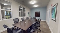 This single-floor professional meeting space is designed to facilitate focused collaboration and productive gatherings. The room centers around a substantial dark wood conference table surrounded by ergonomic mesh-back chairs, comfortably accommodating a sizable group for meetings, presentations, or discussions. The walls are finished in a calming combination of light gray and soft blue, lending the space a composed, professional atmosphere. Flush-mounted circular ceiling fixtures provide consistent, even illumination throughout the room. A large wall-mounted display screen serves as the focal point for presentations and video conferencing, positioned prominently for clear sightlines from all seating positions. A framed piece of artwork adds a touch of personality to the otherwise functional interior. The room features a large interior window, allowing borrowed light from an adjacent space while maintaining privacy. Patterned carpet tiles cover the floor, adding texture and contributing to the room's acoustic qualities. Subtle decorative accents on the table, including a modern circular ornament, complete the polished yet approachable aesthetic of this well-appointed meeting space.