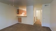 This single-floor residence offers a practical and functional layout designed for comfortable everyday living. The living area is open and carpeted, providing a warm and inviting atmosphere that flows seamlessly toward the kitchen. The kitchen features a pass-through counter with a raised breakfast bar, warm honey-toned wood cabinetry above, and under-counter storage, creating an efficient and connected cooking space that remains open to the main living area. A textured ceiling adds character throughout the space. A central hallway extends from the living area, leading to additional rooms beyond, suggesting a well-organized floor plan with clear separation between living and private spaces. The neutral, light-colored walls throughout lend a clean and airy feel, while the carpeted floors add softness underfoot. The layout makes efficient use of the available space, with thoughtful transitions between areas. Overall, this residence presents a straightforward and comfortable living environment with a cohesive design that balances openness in the common areas with accessible private spaces down the hall.