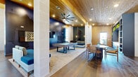This generously sized single-floor space is designed as a sophisticated amenity and communal hub, thoughtfully organized across a living room, a dedicated office, and two additional rooms. Throughout the space, warm natural wood plank ceilings add architectural character and a welcoming warmth, complemented by wide-plank hardwood flooring that flows continuously from room to room.

The expansive living room serves as the social centerpiece, featuring a pool table atop a large area rug, comfortable lounge seating, and a dramatic dark accent wall. Large ceiling fans and recessed lighting keep the atmosphere well-lit and comfortable. Built-in navy shelving units line one wall, styled with greenery and decorative accessories, while vertical wood paneling adds textural depth.

The office is enclosed in sleek steel-framed glass partitions, offering transparency while maintaining function. Inside, a conference table and task chairs are positioned beneath the wood ceiling, with floor-to-ceiling windows providing abundant natural light and outward views.

Additional areas include a games room with a shuffleboard table and high-top seating, and corridors lined with light-toned stone tile walls. Restroom facilities are also present. Together, the varied zones create a dynamic, design-forward environment suited for both productivity and leisure.