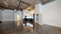 Located at 11135 Weddington St in Los Angeles, this single-floor industrial-modern loft offers a striking open-plan layout defined by soaring ceilings with exposed concrete soffits, visible ductwork, and track lighting that together create an authentic urban-industrial aesthetic. Polished concrete floors run throughout the expansive main area, reflecting light and enhancing the sense of scale. A central structural column anchors the main open space, which is bathed in natural light through a generous bank of large windows along the front facade, offering views of surrounding greenery. The fully equipped kitchen features sleek two-tone cabinetry — dark lower cabinets paired with light uppers — complemented by dark countertops, a patterned tile backseat with under-cabinet LED lighting, a gas range, stainless microwave, refrigerator, and dishwasher, all in a modern black finish palette. A dedicated laundry area with a stacked washer/dryer is conveniently enclosed nearby. A full bathroom includes a backlit vanity mirror, dark-topped vanity, and polished concrete flooring consistent with the rest of the space. Additional rooms or flex spaces are accessible through interior doorways, offering versatile utility. The overall ambiance is clean, open, and contemporary, well-suited for creative, live-work, or professional uses.