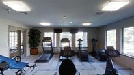This fitness center offers a functional and inviting workout environment designed to support a variety of cardio-focused exercise routines. The room is outfitted with multiple treadmills, elliptical trainers, and a stair climber, all arranged to face a bank of large windows that line the far wall, allowing generous natural light to fill the space and providing views of the surrounding greenery outdoors. Mounted flat-screen televisions are positioned above the windows, giving users entertainment options during their workouts. The flooring features a patterned carpet with light blue and gray tones, adding a modern aesthetic to the room. Recessed lighting and flush-mounted ceiling fixtures supplement the natural light, ensuring the space remains well-lit at all times. Neutral-toned walls contribute to a calm, focused atmosphere. A decorative potted tree in the corner adds a touch of greenery to the interior, softening the utilitarian nature of the space. The layout is open and organized, providing comfortable spacing between each piece of equipment for ease of movement and use.