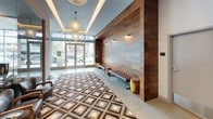 This contemporary lobby makes a striking first impression through its thoughtful blend of warm and modern design elements. A full-height wood-paneled feature wall serves as the centerpiece, its rich walnut tones adding warmth and texture against the cooler tones of the polished concrete flooring. A bold geometric patterned area rug in earth tones anchors the seating area, where tufted leather benches and accent stools invite residents and guests to pause.

Natural light flows generously through expansive floor-to-ceiling glass panels flanking the main entry doors, offering clear views of the surrounding streetscape and connecting the interior to the outdoors. Overhead, a coffered ceiling detail frames a decorative globe pendant light, with recessed cove lighting adding a soft ambient glow throughout.

Adjacent to the main entry area, glass-enclosed rooms provide dedicated spaces for a concierge or management desk, complete with built-in wood shelving and workstations. A corridor extends deeper into the building, lined with additional pendant lighting in varied warm tones, leading to a lounge area furnished with casual seating and round tables. A stainless steel elevator provides vertical access, seamlessly integrated into the lobby's refined aesthetic. The overall ambiance is modern, curated, and welcoming.