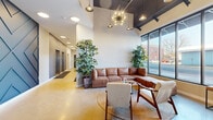 This well-appointed residential lobby offers a thoughtfully designed, single-floor entry experience that blends industrial and contemporary aesthetics. Polished concrete floors span the entire space, reflecting natural light that pours in through expansive floor-to-ceiling windows and black-framed glass storefront walls facing the street. The soaring ceilings feature exposed ductwork and dark-painted structural elements, complemented by a curated mix of pendant lighting — including a striking sputnik chandelier and Edison-style bulb fixtures — that add warmth and character throughout.

The lounge area is furnished with a brown leather sectional sofa, mid-century modern accent chairs, and a woven area rug, all anchored by tall indoor trees that introduce a natural, organic element to the space. A bold geometric accent wall in deep slate blue frames the elevator bank, creating a dramatic focal point along the corridor. Adjacent to the lounge, a dedicated mailbox station features a built-in bank of individual metal mailboxes set into a clean white partition wall, ensuring practical functionality without compromising the lobby's polished look.

The entry vestibule, framed by black-framed glass partitions and doors, provides a seamless transition from the exterior, flooding the space with additional daylight. Overall, the lobby delivers a welcoming, modern atmosphere with strong architectural detail and high-end finishes throughout.