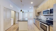 This well-appointed single-floor residence showcases a contemporary design with a thoughtful layout throughout. The open-concept kitchen is a standout feature, boasting light wood cabinetry, dark granite countertops, and a striking herringbone-patterned tile backsplash. A central island with matching granite provides additional prep space and casual seating, illuminated by a pair of stylish black-and-gold pendant lights. Stainless steel appliances, including a French door refrigerator, electric range, and built-in microwave, complete the kitchen's modern aesthetic. Light-toned hardwood-style flooring flows seamlessly from the kitchen into the open living area, which benefits from natural light through multiple windows and is finished with a ceiling fan overhead. A dedicated in-unit laundry area is conveniently accessible from the kitchen. The bedrooms are finished with plush carpeting and ceiling fans, offering a comfortable retreat, with natural light filtering through window blinds. The primary bedroom connects to a spacious walk-in closet featuring built-in shelving and clothing rods. The bathroom is refined in design, featuring light wood vanity cabinetry, dark granite countertops, a backlit mirror, and a glass-enclosed shower with large-format marble-look tile. Throughout, the space maintains a clean, neutral palette that is both modern and inviting.