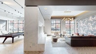 This sophisticated single-floor amenity space is designed with a contemporary aesthetic, combining high-end materials and curated furnishings throughout its open, flowing layout. The herringbone-patterned hardwood flooring serves as a unifying element across the entire space, lending warmth and visual interest underfoot.

A striking marble-clad fireplace anchors one section of the lounge, providing an elegant focal point alongside a seating area furnished with a large leather sofa, accent chairs, and gold-framed coffee tables. A bold floral wallcovering adds a dramatic decorative touch, while an angular, sculptural gold chandelier suspended from a recessed tray ceiling introduces refined ambient lighting. Floor-to-ceiling black-framed windows flood the lounge with natural light and offer views of the surrounding streetscape.

The kitchen area features a generously sized island with dramatic veined granite countertops, complemented by bold black pendant lights with gold interiors and green velvet barstools. Stainless steel appliances, textured dark tile backsplash, and brass fixtures complete the polished culinary space.

Adjacent to the kitchen, a game room houses a pool table and foosball table, offering a casual and social counterpoint to the more refined lounge areas. A cozy nook with a mustard yellow banquette provides an intimate retreat within the broader open-plan environment.