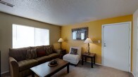 This single-floor space centers around a well-appointed living room designed for comfort and functionality. The room features warm yellow and tan accent walls that create an inviting atmosphere, complemented by neutral-toned carpeting that extends throughout. A textured popcorn ceiling adds a classic architectural element to the space.

The living area is furnished with a dark brown sofa adorned with patterned and solid throw pillows, paired with a light-colored armless accent chair, offering flexible seating arrangements. A dark wood rectangular coffee table anchors the seating area, while two matching wrought-iron table lamps provide warm ambient lighting on either side of the room.

A framed wall mirror positioned above the accent chair reflects light and adds depth to the space. A large window along one wall allows natural light to filter into the room, brightening the interior. The entryway door, finished in white, is conveniently located within the living area. Overall, the space presents a cozy, livable environment with a cohesive color palette and practical layout.