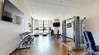 Located at 2142 Commonwealth Ave in Charlotte's 28205 zip code, this fitness center is a well-appointed, single-floor facility designed to support a comprehensive workout experience. The space features polished concrete flooring throughout, lending it a clean, durable, and modern aesthetic that is easy to maintain.

The main workout area is generously sized and filled with natural light, thanks to large, multi-pane windows along one wall that provide views of the outdoors. A wall-mounted flat-screen television is positioned for easy viewing from the cardio section, which includes multiple treadmills and an elliptical machine. A full rack of dumbbells lines one wall, offering a wide range of weight options for free weight training.

The facility also includes a multi-functional cable machine system, an incline bench, and a selection of resistance bands and TRX-style suspension training equipment mounted near the windows. Stability and exercise balls are stored in a dedicated area, providing options for core and flexibility training.

A secondary area connects to the main workout room and features a dual-level water fountain station, recessed lighting, and drop tile ceilings throughout. The entryway is marked with a branded floor mat and provides direct access through glass-paned doors. The overall layout is open and functional, offering users a full range of cardio, strength, and flexibility training options in one cohesive space.