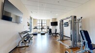 Located at 2142 Commonwealth Ave in Charlotte's 28205 zip code, this fitness center is a well-appointed, single-floor facility designed to support a comprehensive workout experience. The space features polished concrete flooring throughout, lending it a clean, durable, and modern aesthetic that is easy to maintain.

The main workout area is generously sized and filled with natural light, thanks to large, multi-pane windows along one wall that provide views of the outdoors. A wall-mounted flat-screen television is positioned for easy viewing from the cardio section, which includes multiple treadmills and an elliptical machine. A full rack of dumbbells lines one wall, offering a wide range of weight options for free weight training.

The facility also includes a multi-functional cable machine system, an incline bench, and a selection of resistance bands and TRX-style suspension training equipment mounted near the windows. Stability and exercise balls are stored in a dedicated area, providing options for core and flexibility training.

A secondary area connects to the main workout room and features a dual-level water fountain station, recessed lighting, and drop tile ceilings throughout. The entryway is marked with a branded floor mat and provides direct access through glass-paned doors. The overall layout is open and functional, offering users a full range of cardio, strength, and flexibility training options in one cohesive space.