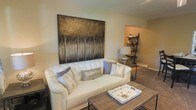 This single-floor residence offers a thoughtfully designed living space that balances comfort and contemporary style. The living room serves as the focal point of the home, anchored by a plush tufted white sofa adorned with coordinating metallic and neutral accent pillows. A large-scale nature-inspired artwork — depicting a stand of trees in rich golden and dark tones — commands attention on the main wall, adding depth and visual drama to the space. The warm beige and taupe color palette creates a cohesive, calming ambiance throughout. A rustic wood coffee table with a decorative tray adds texture and a grounded, earthy contrast to the lighter furnishings. Adjacent to the main living area, an open nook or secondary space is visible, featuring a lean-to shelving unit that provides practical display and storage options, alongside a table lamp with a mirrored base that contributes a touch of elegance. The layout flows naturally between areas, making the space feel open and connected. Neutral-toned walls and warm flooring tie the rooms together, while the overall design aesthetic leans toward modern transitional — sophisticated yet approachable. This residence is well-suited for those seeking a stylish, move-in-ready home with a warm and welcoming character.