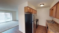 This single-floor space features a compact, functional kitchen equipped with stainless steel appliances, including a refrigerator and electric range. Oak-toned upper cabinetry is mounted above the refrigerator, providing additional storage. The kitchen showcases durable hardwood-style flooring, which contrasts with the carpeted living area visible through the open passageway. A textured ceiling and neutral wall tones throughout give the space a clean, understated appearance. The layout connects the kitchen directly to the adjacent living area, promoting an open flow between spaces. A baseboard heater is visible along the living area wall, indicating an electric heating system. Flush-mount ceiling lighting provides adequate illumination in the kitchen area. The overall design is practical and straightforward, prioritizing functionality with modest finishes suited for everyday living.
