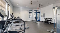 Located in Oviedo, Florida, this fitness center offers a well-appointed, single-floor workout space designed to support a comprehensive exercise routine. The interior is finished with crisp white walls accented by bold blue window and door frames, creating a clean, energizing aesthetic throughout. Durable rubber flooring covers the entire space, providing cushioning and traction suited for a variety of workouts.

The cardio zone is anchored by multiple TRUE-brand treadmills and an elliptical trainer, positioned in front of large arched and framed windows that allow generous natural light and outdoor views to enhance the workout experience. Ceiling fans and recessed lighting ensure the space remains comfortable and well-lit at all times.

The strength training area features a multi-station cable machine set against a bold blue accent wall, a tiered dumbbell rack with a full range of free weights, and an adjustable weight bench, offering options for both functional and resistance training. A wall-mounted flat-screen television overlooks the strength zone, adding an entertainment element to training sessions.

Full-length mirrors are strategically placed to allow users to monitor their form. Multiple entry and exit points, including glass-paneled blue doors, connect the space to the surrounding outdoor environment, reinforcing an open, accessible feel throughout this thoughtfully equipped fitness facility.