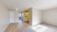 This single-floor residence offers a practical and well-organized layout, encompassing a combined living, dining, and kitchen area alongside one bedroom, one bathroom, and two closets. The open-concept main living area features soft carpet flooring, neutral beige walls, a textured ceiling, and a ceiling fan with light fixture. A wall-mounted HVAC unit and a window with blinds provide climate control and natural light, while a front entry door with security hardware anchors the space. The kitchen is separated from the living area by a partial wall and features light hardwood flooring, white cabinetry with ample upper and lower storage, white countertops, a double basin sink, and a full suite of white appliances including a refrigerator, electric range, built-in microwave, and dishwasher. The bedroom is generously sized, carpeted, and includes a ceiling fan with lighting, a window for natural light, and access to a walk-in closet with rod and shelving. A second closet provides additional storage elsewhere in the unit. The consistent use of neutral tones, textured ceilings, and white trim throughout creates a cohesive, clean aesthetic that maximizes the functionality of the overall floor plan.