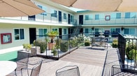 This two-story multi-unit property is thoughtfully designed around a vibrant central courtyard that serves as the heart of the complex. The exterior facade is finished in warm yellow stucco, accented by black iron railings that line the upper-level balconies and walkways, lending the building a clean, classic character.

The landscaped courtyard is a standout feature, filled with lush tropical plantings including bird-of-paradise, climbing roses, and a variety of flowering shrubs. A tiered decorative stone fountain anchors one end of the courtyard, adding an elegant focal point and a sense of serenity to the outdoor space. Ground-level corridors run along the building's interior wings, finished with green turf walkways that connect individual units and provide easy outdoor access throughout.

A generous wooden deck extends from one section of the property, furnished with iron seating and enclosed by black metal railings. This area is shaded by sail canopies, creating a comfortable outdoor gathering space that overlooks the courtyard garden below. An exterior staircase connects the ground and upper levels, and utility storage is accessible via a green metal structure on the deck level.

The upper-level balconies offer private outdoor space for each unit, with views over the landscaped grounds. Ample natural light reaches all areas of the property thanks to the open courtyard design, making this a bright and inviting community-oriented space.