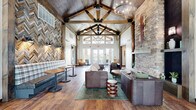 Located at 1810 NW Savannah Ln in Ankeny, Iowa, this single-floor community clubhouse offers a generously sized, well-appointed gathering space designed to serve a wide range of recreational and social needs. The centerpiece of the facility is a striking great room featuring soaring vaulted ceilings with exposed wooden beams, a herringbone-patterned reclaimed wood accent wall, a stacked stone fireplace, and warm hardwood floors throughout. Large windows and glass doors flood the space with natural light while providing views of the surrounding neighborhood. Adjacent living areas include a lounge with a tray ceiling, an elegant chandelier, and a pool table, creating an inviting space for casual recreation. A dedicated game room with high-top seating and a billiards table adds to the entertainment options. The dining area is distinguished by a bold navy accent wall, contemporary artwork, and a stylish multi-bulb pendant light fixture, complemented by large windows that bring in abundant daylight. The expansive fitness center is comprehensively equipped with treadmills, stationary bikes, strength machines, free weights, and a punching bag, all set within a bright, mirror-lined space with ceiling fans and exterior-facing windows. The hallways feature curated artwork, built-in seating, and teal accent walls, maintaining a cohesive and welcoming design aesthetic throughout.