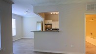 Located at 1300 Knoll Cir in Durham, North Carolina, this single-floor residence offers a clean, modern layout designed for comfortable everyday living. The open-concept living area is finished with light hardwood-style flooring and neutral wall tones, creating a bright and welcoming atmosphere throughout. Crown molding adds a refined architectural detail that runs along the ceiling, lending a polished look to the main living space. The kitchen is accessed via a distinctive pass-through opening framed by a half-wall, which also serves as a breakfast bar with a granite countertop featuring subtle speckled patterning. Inside the kitchen, white upper cabinetry provides ample storage, while a tile backsplash in a warm earth tone adds visual texture behind the stainless steel range. Track lighting above the kitchen workspace ensures the area is well-lit for cooking and meal prep. The layout promotes a natural flow between the kitchen and living areas, making the space feel connected and functional. Large windows allow natural light to filter into the living room, enhancing the airy feel of the interior. The space presents a well-maintained, move-in-ready environment with thoughtful design details throughout.