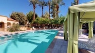 This single-story property showcases a refined Mediterranean-inspired architectural style, characterized by stucco exteriors, arched windows, and a graceful colonnade walkway with stone-tiled flooring. The covered corridor runs the length of the building, framed by robust square columns with decorative detailing and arched openings that frame views of the beautifully landscaped grounds beyond.

The outdoor amenities are a true highlight of the space. A generously sized swimming pool takes center stage, its crystal-clear turquoise water surrounded by travertine tile decking. Poolside loungers are arranged throughout the deck, complemented by stylish green fabric cabanas that provide shaded retreats for relaxation. Towering date palms and mature shade trees envelop the pool area, creating a lush, resort-like atmosphere under brilliant blue skies.

The courtyard area features a decorative fountain surrounded by manicured landscaping, including colorful flowering plants, desert-adapted greenery, and additional palm trees, all set against a backdrop of wide stone pathways. The overall ambiance of the property is one of sun-soaked serenity, blending classic architectural elegance with thoughtfully designed outdoor living spaces that invite leisure and relaxation.