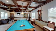 This billiards room is a thoughtfully designed recreational space that blends classic craftsmanship with modern amenities. The most distinctive architectural feature is the rich, dark wood coffered ceiling that spans the entire area, adding depth, warmth, and a refined character to the room. Neutral-toned carpet and light walls complement the dark wood trim and wainscoting that runs throughout, creating a cohesive and inviting atmosphere.

At the heart of the space is a full-size pool table with a vibrant teal felt surface, illuminated by a classic pendant light overhead. A wall-mounted cue rack keeps equipment neatly organized, while bar-height seating along the walls provides convenient spots for spectators. Natural light enters through multiple windows fitted with blinds, and French doors offer access to an outdoor area.

Adjacent to the billiards area is a kitchenette featuring dark cabinetry, granite countertops, and bar stool seating — ideal for refreshments during gatherings. A separate seating nook with a round dining table, upholstered chairs, and a wall-mounted television offers a space for more relaxed socializing. A lounge area with cushioned armchairs rounds out the room's versatile layout, making this space well-suited for both casual recreation and group entertainment.