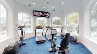 This fitness center is designed to provide an energizing and comfortable workout environment. The space is anchored by a collection of modern cardio equipment, including treadmills, stationary bikes, and elliptical machines, all positioned to take full advantage of the room's standout architectural feature: a series of large, elegantly arched windows. These windows flood the interior with abundant natural light and offer direct views of a sparkling outdoor pool and lush surrounding greenery, creating a refreshing backdrop for any workout.

The walls are painted a clean, crisp white, enhancing the bright and open atmosphere throughout the space. Underfoot, vivid blue carpet tiles add a bold pop of color while providing a comfortable, cushioned surface. Overhead, a ceiling fan ensures steady air circulation, complementing the room's comfortable climate. A wide, wall-mounted flat-screen TV display is positioned prominently above the equipment, allowing users to stay entertained during their sessions. The overall ambiance is modern, well-lit, and inviting, combining functional fitness amenities with an aesthetically polished design.