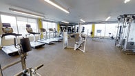 This fitness center offers a comprehensive workout environment designed to accommodate a range of exercise needs. The space is anchored by a row of treadmills and elliptical machines positioned along a bank of large windows, allowing natural light to fill the room while providing views of the surrounding area. Overhead, linear fluorescent lighting ensures the space remains well-lit throughout. The flooring consists of durable rubber-tiled material, providing both safety and comfort underfoot during workouts. A selection of strength training equipment, including cable machines and resistance apparatus, is distributed throughout the floor, offering versatility for users at different fitness levels. Mirrored walls along one side visually expand the space and allow users to monitor their form during exercise. Yellow accent details on select equipment add a subtle pop of color against the neutral grey and white palette of the room. Exposed ductwork along the ceiling contributes an industrial aesthetic to the otherwise clean, modern interior. The overall layout is open and uncluttered, promoting ease of movement between stations and creating an accessible, functional fitness environment.