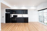 This single-floor space presents a thoughtfully designed open-concept layout where the kitchen flows seamlessly into the living area. The kitchen is a standout feature, outfitted with deep navy shaker-style cabinetry accented by gold hardware, a white subway tile backsplash, light quartz countertops, a stainless undermount sink with a matte black faucet, and black appliances including a range and built-in microwave. Upper cabinets incorporate glass-panel inserts, adding visual depth.

Warm-toned hardwood flooring runs throughout the entire unit, creating a cohesive and inviting feel. The open living area is bright and generously proportioned, benefiting from multiple large windows and a glass-paned balcony door that invite natural light and provide views of the surrounding urban landscape. The balcony access enhances the sense of openness and connection to the outdoors.

A dedicated bedroom area includes convenient in-unit laundry with a stacked washer and dryer tucked neatly into a closet. The bathroom is finished with a dark-stained vanity, gold hardware, a white quartz countertop, a matte black faucet, and a framed mirror. Recessed lighting and clean white walls contribute to the space's polished, contemporary aesthetic throughout.