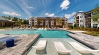 Located in Savannah, Georgia, this outdoor amenity space offers a resort-inspired pool experience designed for relaxation and recreation. The centerpiece is a generously sized swimming pool with crystal-clear water, featuring in-water lounge chairs that allow residents to recline directly in the shallow end — a signature leisure touch. An accessible pool lift ensures the space is welcoming to all. Surrounding the pool, a wide stone-paver deck provides ample room for rows of cushioned chaise loungers and navy blue shade umbrellas, creating a comfortable and well-appointed sunning area. Thoughtfully placed seating vignettes with stylish outdoor furniture offer additional spots to unwind. Lush, manicured landscaping with ornamental shrubs, flowering plants, and mature trees frames the space, adding greenery and privacy. A clubhouse structure with a standing-seam metal roof and covered outdoor patio sits adjacent to the pool, serving as a functional and architectural anchor to the amenity area. The multi-story residential buildings surrounding the space feature contemporary white and gray facades with private balconies that overlook the pool courtyard, blending community living with upscale outdoor amenities beneath the open Georgia sky.