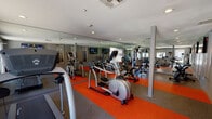 This fitness center is designed to support a comprehensive workout routine within a single, open-plan space. The room is finished with a combination of gray rubberized flooring and bold orange accent strips that visually define the cardio zones and add energy to the environment. Recessed lighting throughout the ceiling ensures the space is well-lit, while wall-mounted flat-screen televisions provide entertainment during workouts.

Floor-to-ceiling mirrors line multiple walls, creating a sense of openness and allowing users to monitor their form. The cardio equipment selection includes elliptical trainers, stationary upright and cycling bikes, and a treadmill, offering a range of low-impact and high-intensity options. A multi-station strength training machine is also present, catering to resistance and weight training needs.

Natural light filters in through several windows and a glass-panel door, which opens to an outdoor area, enhancing the overall atmosphere of the space. The gray-toned walls provide a clean, modern backdrop that complements the equipment and flooring. The layout is thoughtfully organized to allow free movement between stations. Overall, this fitness center provides a functional, well-appointed environment suited for diverse exercise routines.