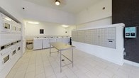 This single-floor shared amenity space serves as both a laundry facility and mail center, designed with functionality and cleanliness in mind. The interior is finished with large-format white floor tiles and crisp white walls, creating a bright, open atmosphere enhanced by overhead lighting and a ceiling fan.

The laundry area is well-equipped with multiple stacked dryer units along one wall and a row of top-loading washing machines along another, providing ample capacity for residents. A central stainless steel utility table with a lower shelf offers a practical surface for sorting and folding laundry.

Adjacent to the laundry area, an extensive bank of individual aluminum mailboxes lines the wall, accommodating a large number of residents. The layout ensures easy access and efficient use of the shared space.

The facility connects directly to what appears to be a communal outdoor area, accessible through full-height glass doors and floor-to-ceiling glazed panels that allow natural light to flood the interior. Through the glass, a furnished outdoor lounge area with colorful seating is visible, suggesting a broader community amenity complex. The transparent entry creates a seamless visual connection between the interior utility space and the vibrant outdoor communal environment beyond.