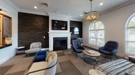 Located at 1155 Museum Blvd in Vernon Hills, Illinois, this single-floor amenity lounge offers a well-appointed social space designed for both relaxation and entertainment. The interior is anchored by a striking fireplace framed by a classic white mantel and flanked by dramatic dark stacked-stone accent walls, creating a warm and inviting focal point. A mix of contemporary lounge chairs, ottomans, and small round coffee tables are arranged to encourage conversation and comfort throughout the space.

Crown molding runs consistently along the ceilings, adding a refined architectural detail that complements the overall design aesthetic. Recessed lighting and elegant drum-style pendant chandeliers provide warm, layered illumination. Tall arched windows along one wall flood the space with natural light and offer views of the surrounding greenery, enhancing the airy ambiance.

A dedicated game area features a large wall-mounted Scrabble board alongside a chalkboard, blue velvet ottomans, and casual seating — ideal for group activities. An electronic dartboard arcade machine adds another entertainment option nearby. The patterned geometric carpeting throughout ties the various zones together cohesively.

A glass-panel door connects to an adjacent corridor, providing easy access through the space. The overall atmosphere balances classic architectural elegance with modern, playful amenities.