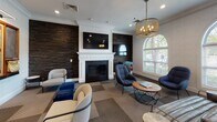 Located at 1155 Museum Blvd in Vernon Hills, Illinois, this single-floor amenity lounge offers a well-appointed social space designed for both relaxation and entertainment. The interior is anchored by a striking fireplace framed by a classic white mantel and flanked by dramatic dark stacked-stone accent walls, creating a warm and inviting focal point. A mix of contemporary lounge chairs, ottomans, and small round coffee tables are arranged to encourage conversation and comfort throughout the space.

Crown molding runs consistently along the ceilings, adding a refined architectural detail that complements the overall design aesthetic. Recessed lighting and elegant drum-style pendant chandeliers provide warm, layered illumination. Tall arched windows along one wall flood the space with natural light and offer views of the surrounding greenery, enhancing the airy ambiance.

A dedicated game area features a large wall-mounted Scrabble board alongside a chalkboard, blue velvet ottomans, and casual seating — ideal for group activities. An electronic dartboard arcade machine adds another entertainment option nearby. The patterned geometric carpeting throughout ties the various zones together cohesively.

A glass-panel door connects to an adjacent corridor, providing easy access through the space. The overall atmosphere balances classic architectural elegance with modern, playful amenities.