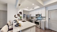 Allura presents a thoughtfully designed single-floor residence that combines contemporary aesthetics with functional living. The open-concept kitchen is a standout feature, equipped with stainless steel appliances including a French door refrigerator, electric range, and built-in microwave. Crisp white cabinetry with sleek hardware lines the walls, complemented by a grey tile backsplash and a generous island with quartz countertops that doubles as a dining and prep space, illuminated by modern pendant lighting.

The living room exudes warmth and character, anchored by a bold teal accent wall that provides a striking backdrop for the space. Wood-look flooring flows throughout, while a ceiling fan and sliding glass door leading to the outdoors enhance comfort and connectivity to the exterior.

Two well-appointed bedrooms offer restful retreats, each featuring distinct accent walls—one in a rich terracotta tone—and large windows that invite natural light. A spacious walk-in closet with wire shelving and ample hanging storage serves the primary suite.

The primary bathroom features a dual-sink vanity with backlit mirrors and grey cabinetry, paired with a glass-enclosed walk-in shower and tile flooring. The secondary bathroom offers a full tub-shower combination with clean tile surround. Recessed lighting and a cohesive neutral palette tie the entire residence together seamlessly.