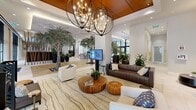 This expansive, single-floor lobby is a masterfully designed communal space that seamlessly blends natural elements with contemporary architecture. The most striking feature is the series of full-grown indoor trees, each planted in illuminated stone-lined floor beds, creating a dramatic biophilic atmosphere that brings the outdoors in. Wide-plank hardwood flooring transitions into large-format porcelain tile, guiding visitors through distinct functional zones. The lounge area is elegantly appointed with a mix of white and rich brown leather sofas, an organic wood coffee table, and layered area rugs, all anchored beneath bold orb-style chandeliers suspended from a warm wood-paneled ceiling tray. A sleek concierge desk, backlit by a textured white stone wall and framed artwork, serves as the welcoming focal point for staff. Nearby, a dedicated mail center features a long, dark-topped bank of numbered mailboxes bathed in natural light from floor-to-ceiling windows. Sculptural wooden screen dividers add warmth and visual interest, while a bar-height counter area provides additional gathering space. Throughout, the design maintains a bright, airy ambiance enhanced by recessed lighting, generous glazing, and lush greenery that together create an inviting, resort-inspired environment.
